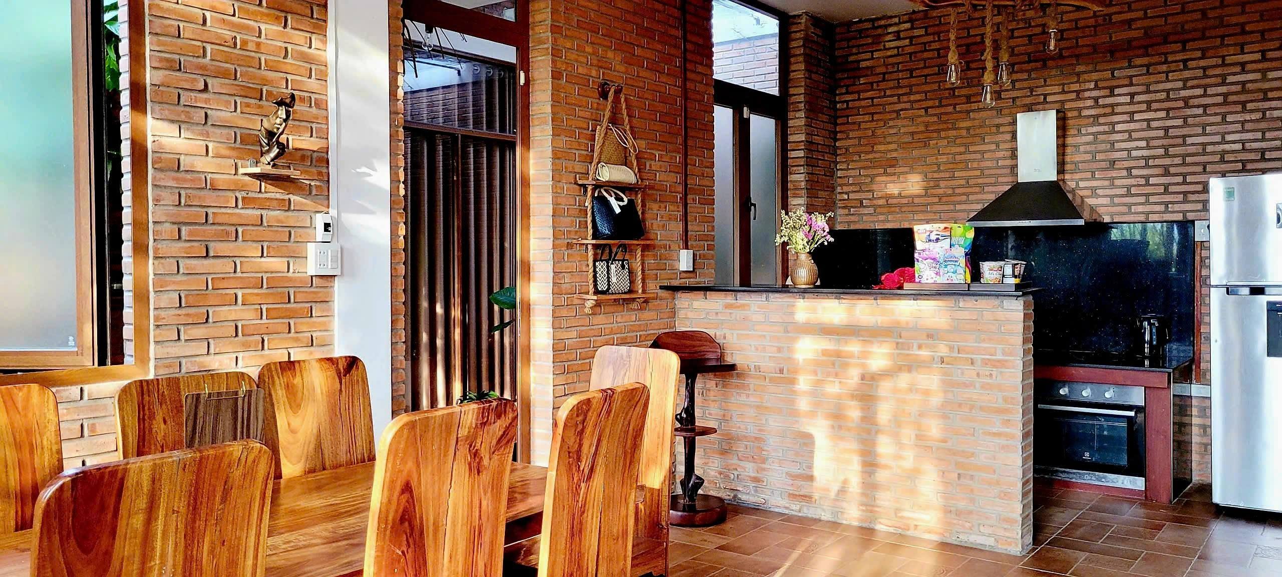 Bếp mở sau quầy gạch với lò nướng, máy hút mùi và tủ lạnh; bàn ăn gỗ nguyên tấm kê phía trước
