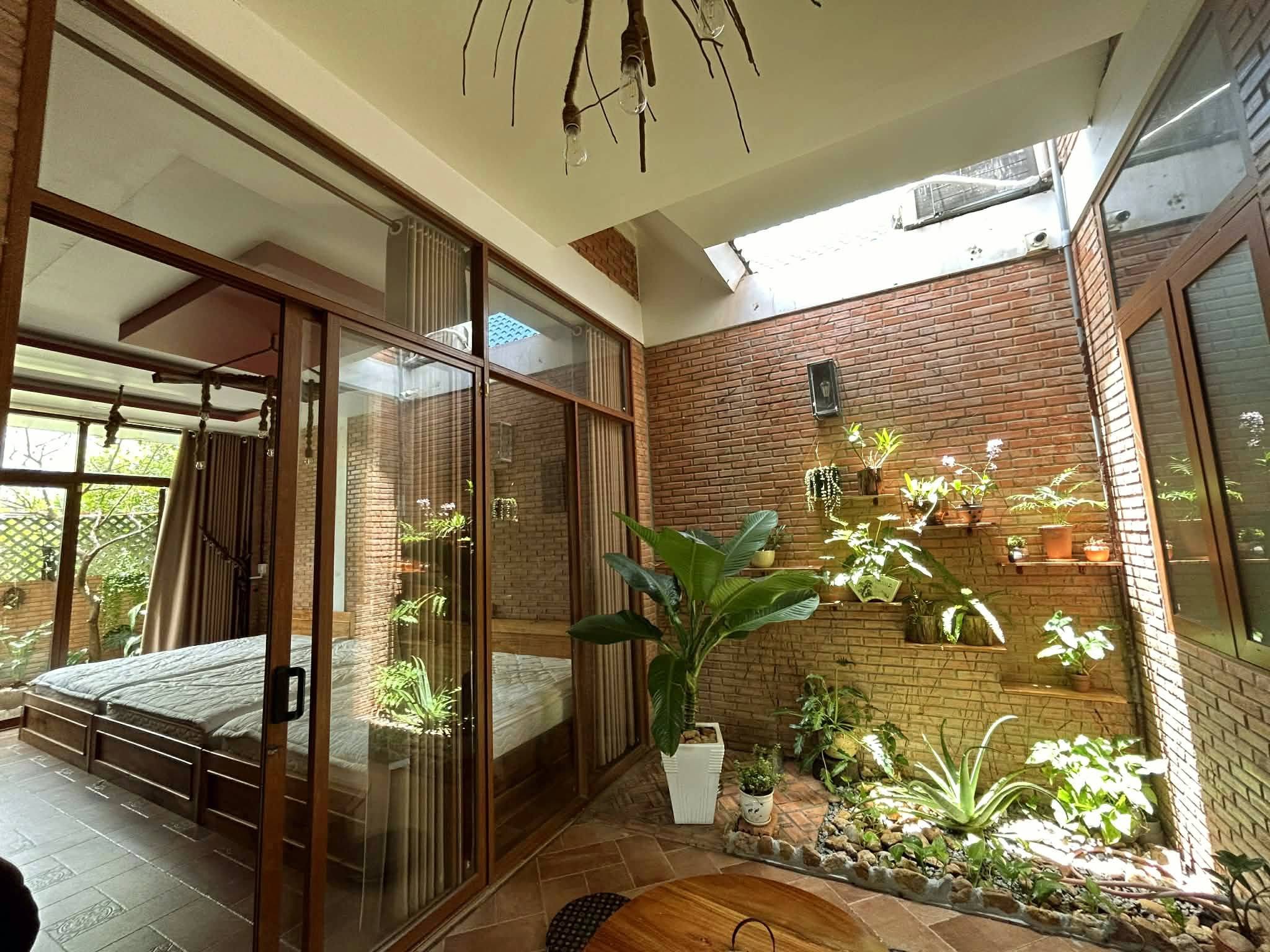 Giếng trời giữa nhà lấy nắng, tường gạch treo kệ gỗ đầy chậu cây, phòng ngủ ngăn bằng vách kính trượt