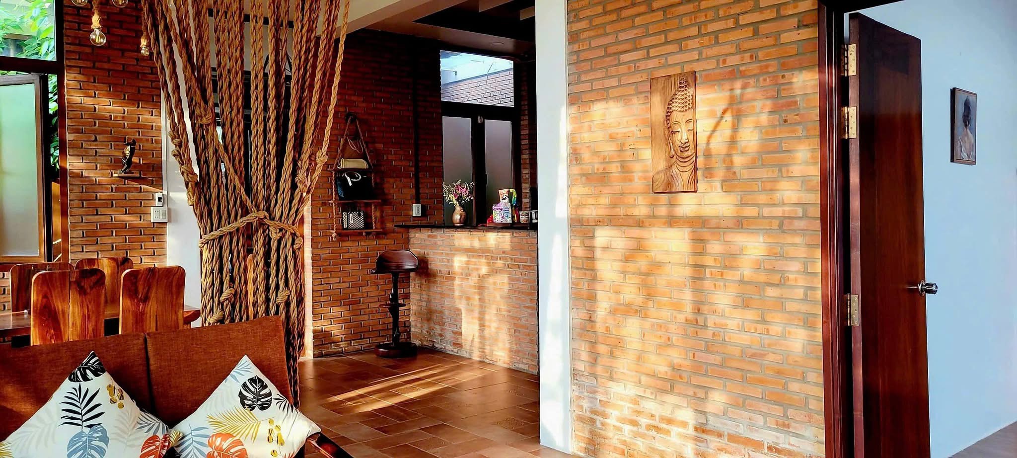 Phòng khách nhìn từ bàn ăn: ghế gỗ nguyên tấm, tường gạch trần treo phù điêu mặt Phật, quầy dẫn vào bếp