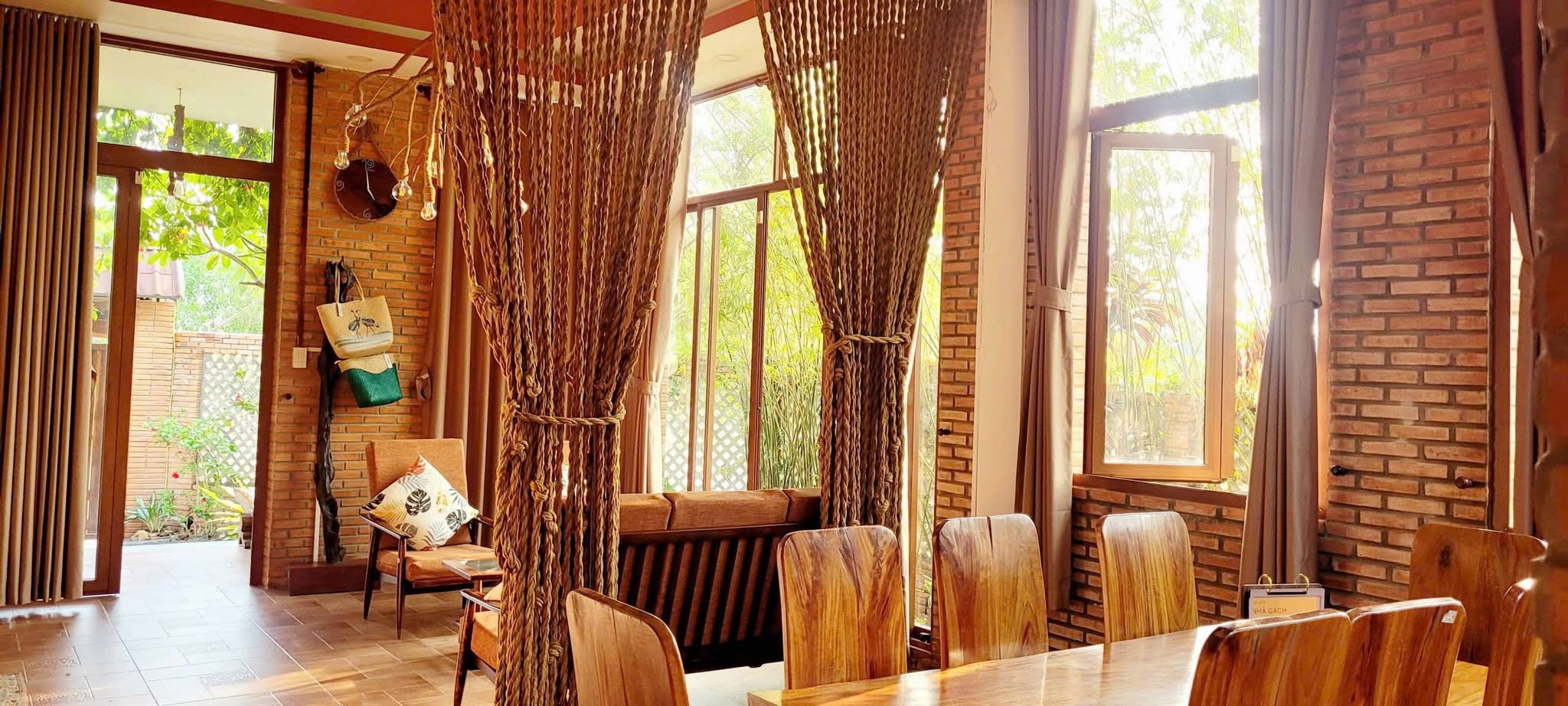 Phòng khách và bàn ăn thông nhau, rèm thừng bện treo giữa hai khu, tường gạch trần và cửa kính cao nhìn ra bụi tre