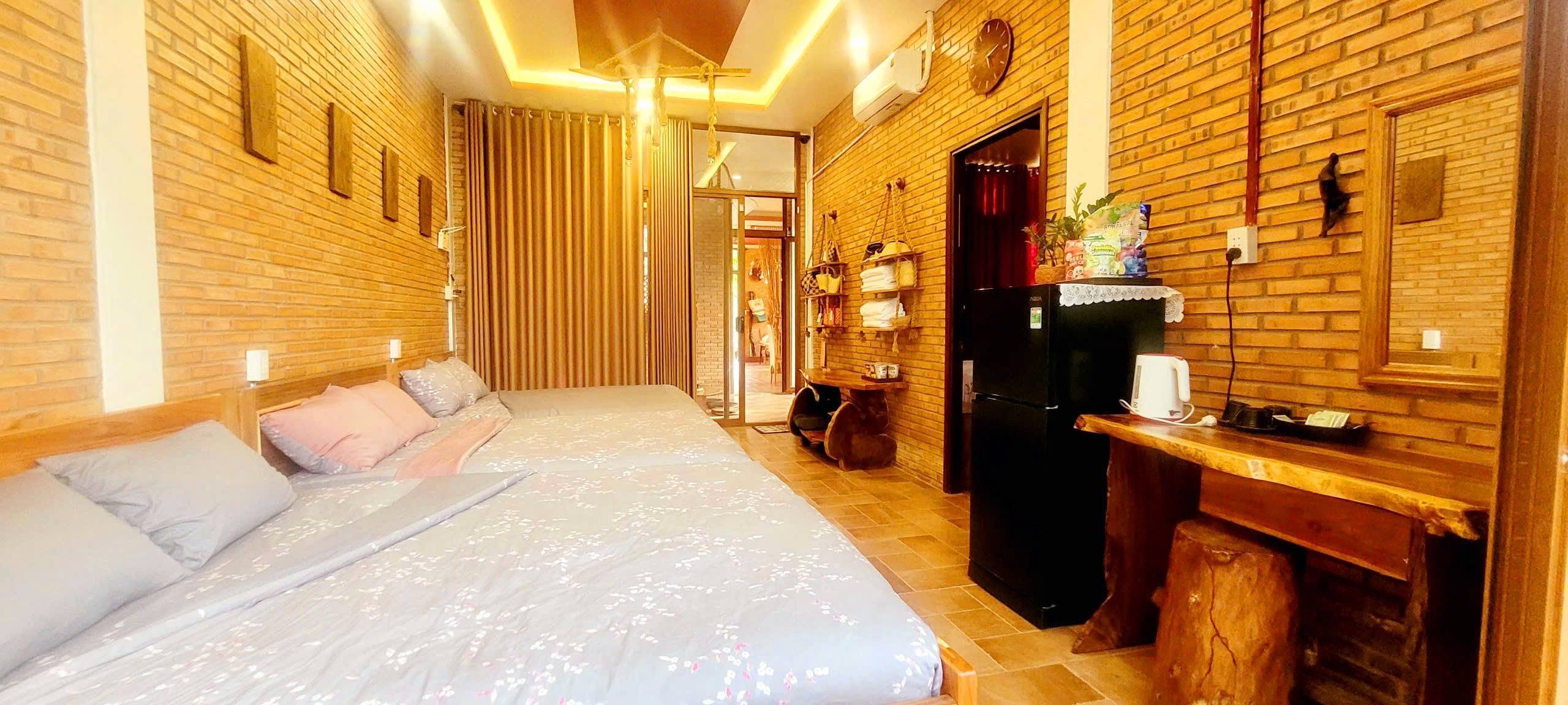 Phòng ngủ tường gạch trần: một giường đôi, tủ lạnh mini, ấm siêu tốc và bàn gỗ nguyên tấm, cửa kính mở ra hành lang